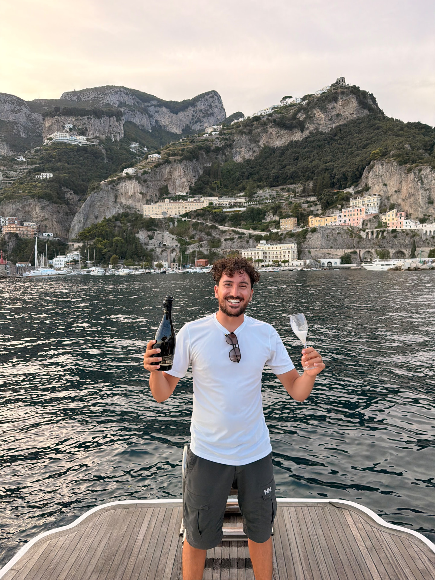 Giovanni with a bottle of prosecco and a glass on board, with Amalfi at sunset behind him